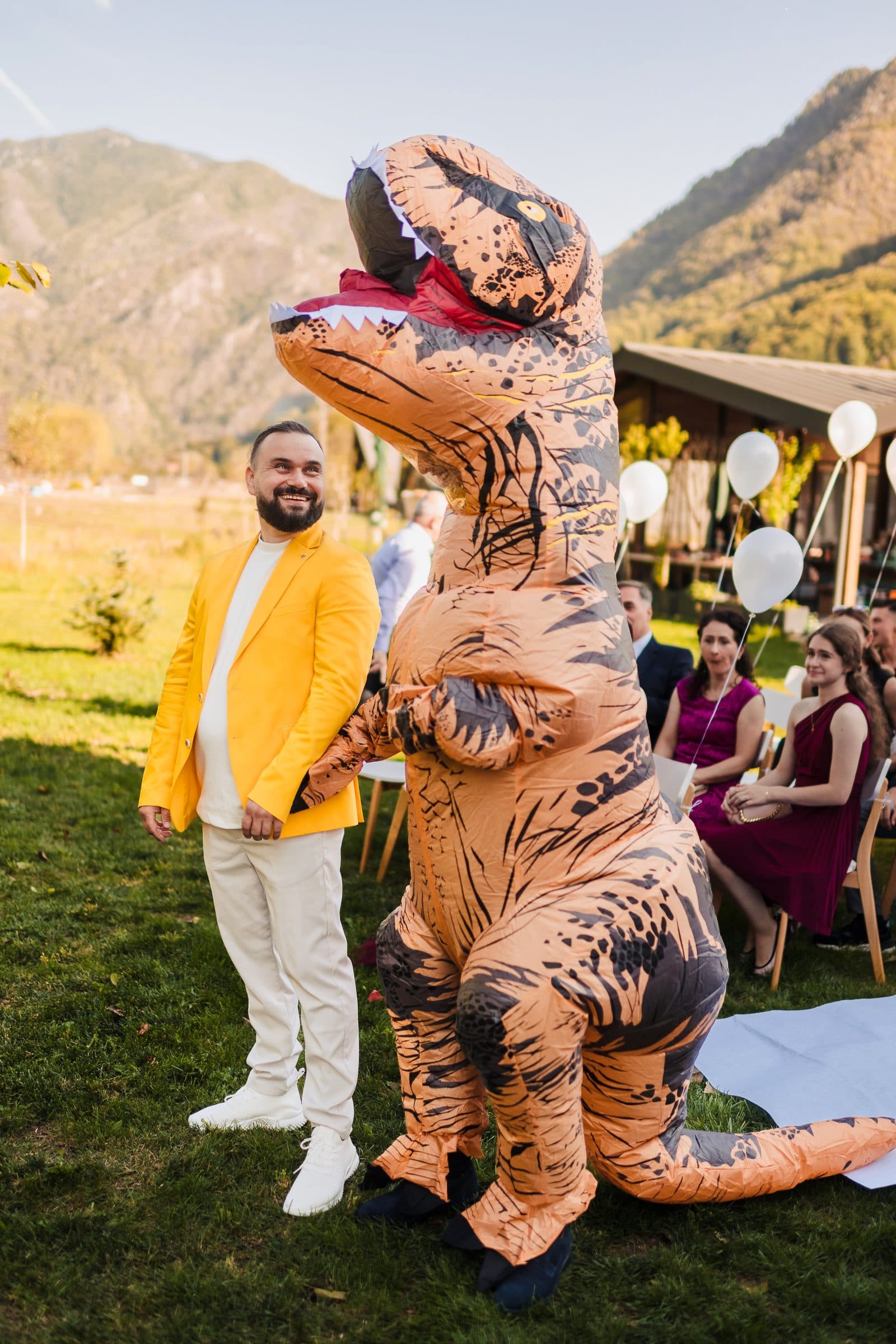 The groom in a yellow jacket waits at the ceremony next to a guest in an inflatable dinosaur costume