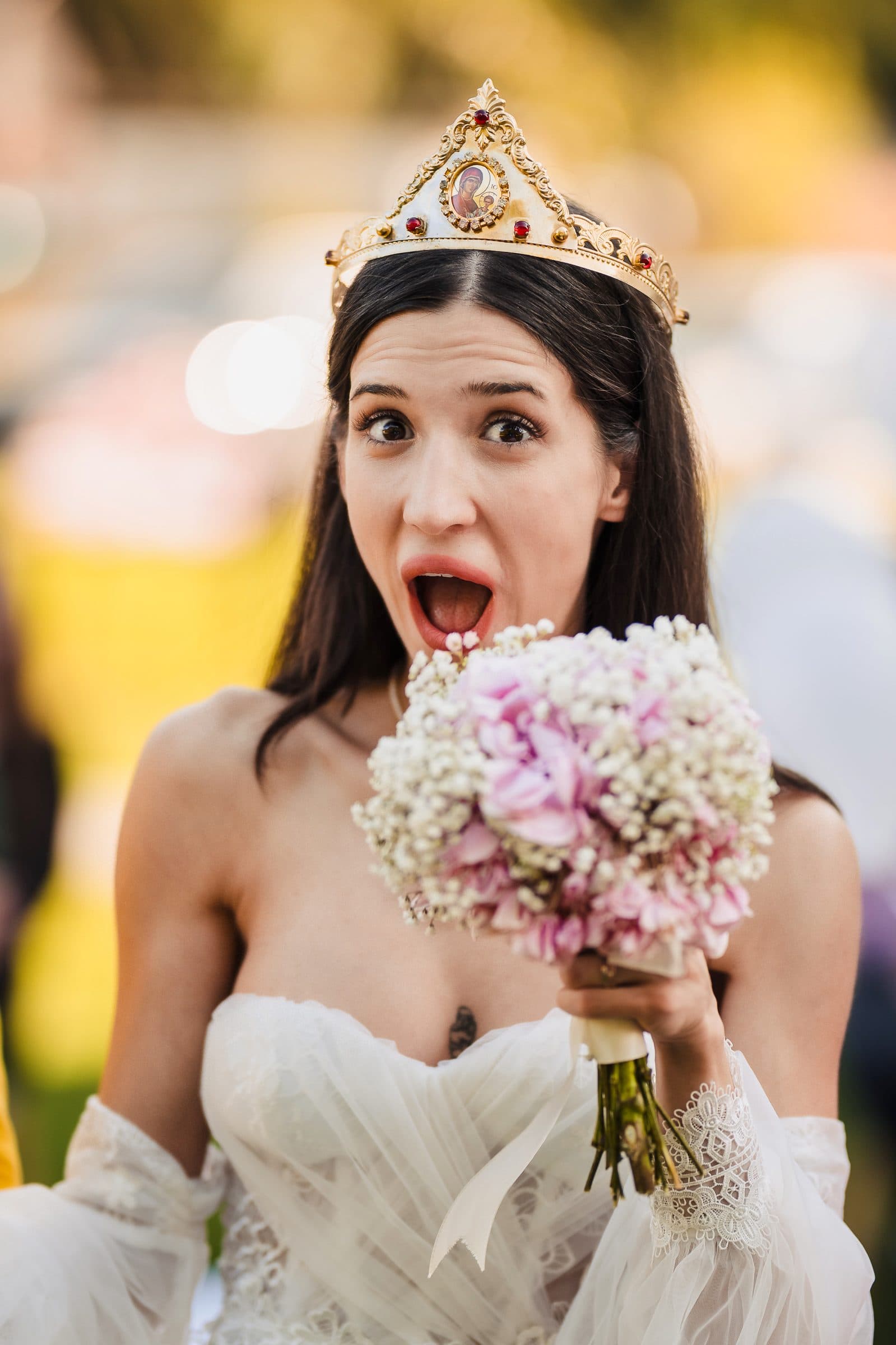 The bride, wearing a wedding crown, pulls a wide-eyed surprised face over her bouquet