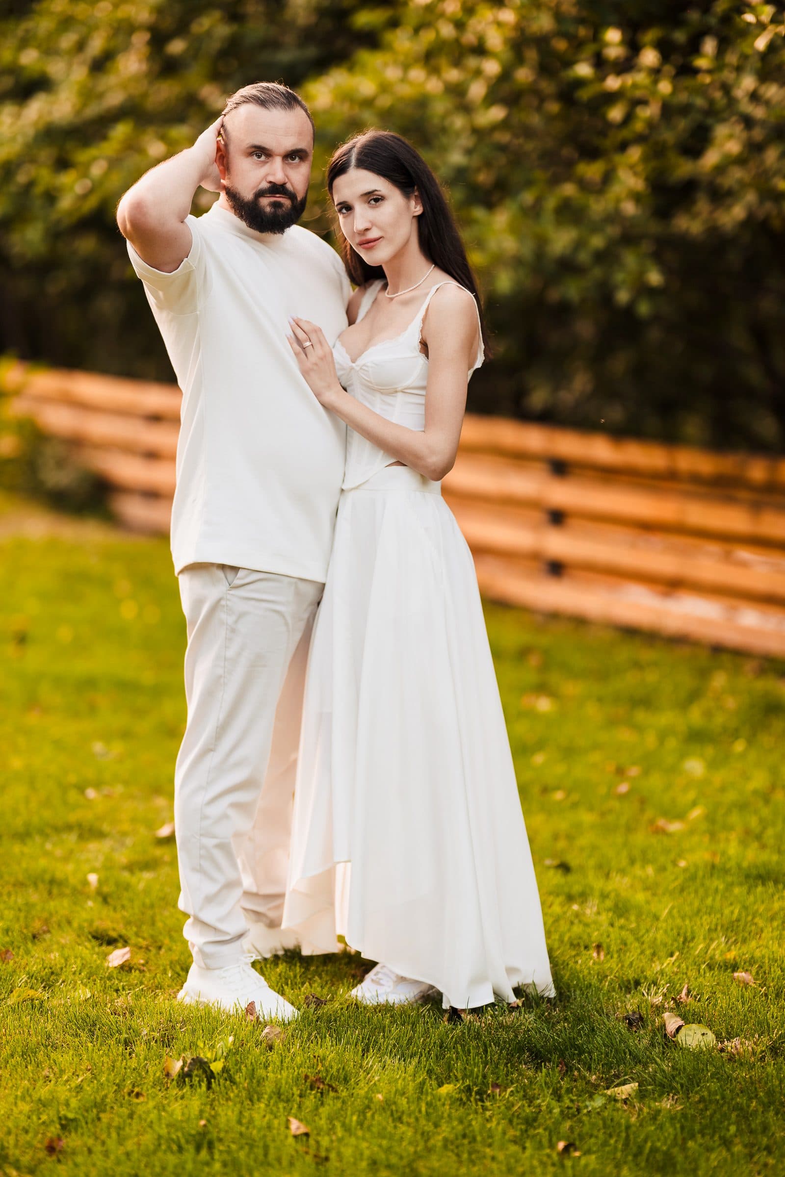 The couple in white on the lawn; he scratches his head, she leans on him