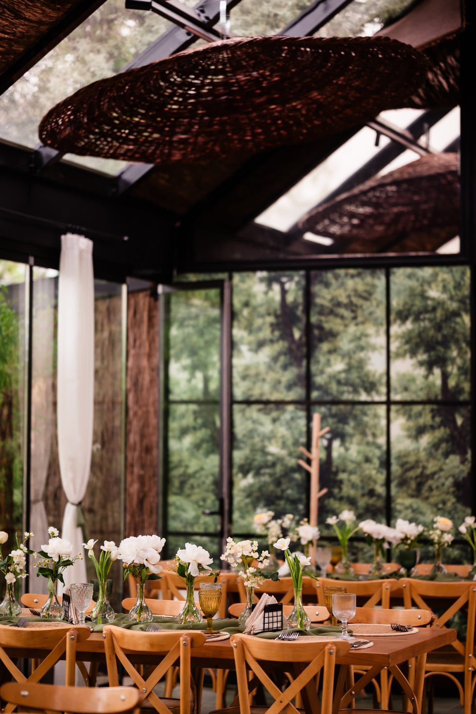Long wooden tables set with white peonies and amber glasses under a woven ceiling