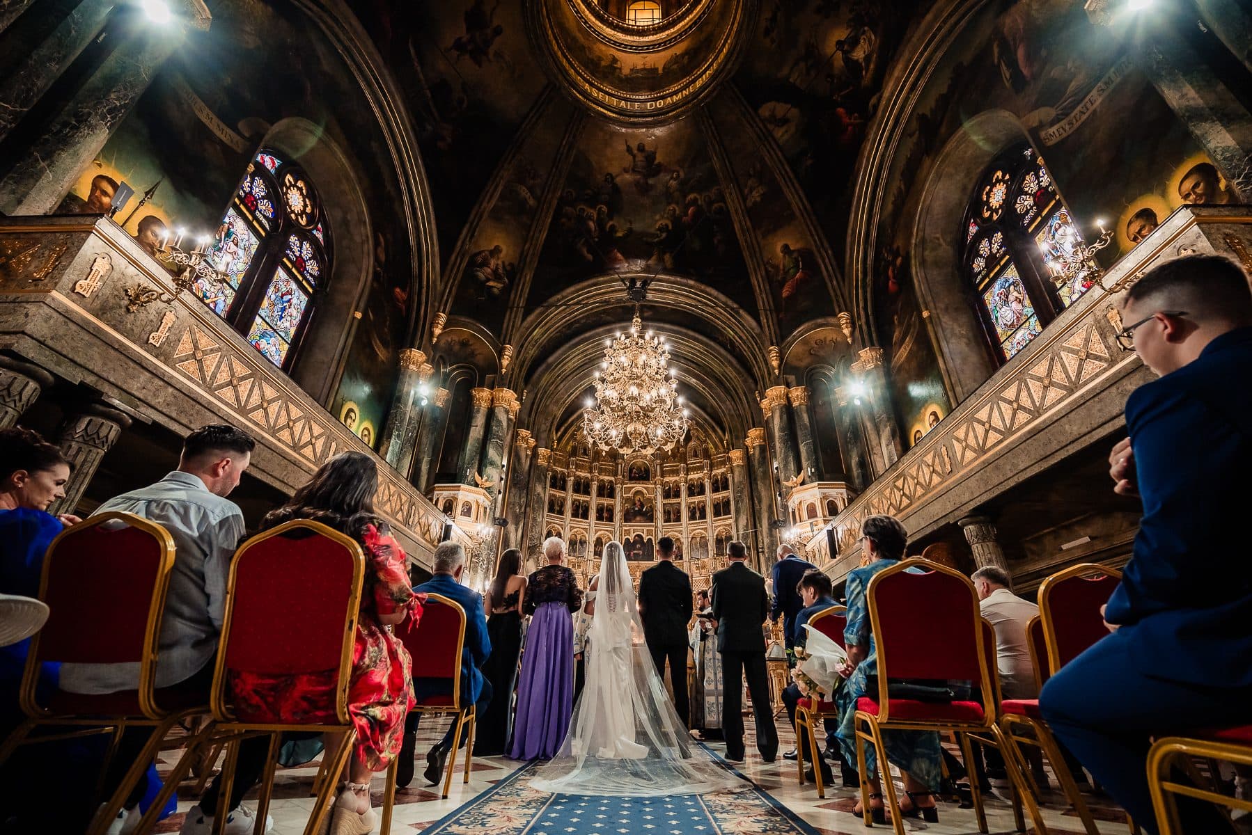 Wide view of the wedding ceremony inside a painted cathedral, guests seated in red chairs
