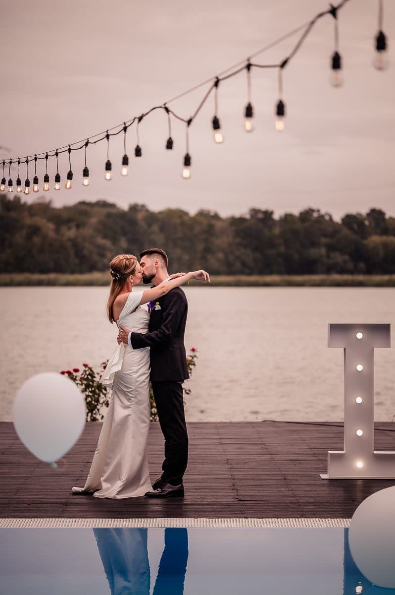 The couple embraces on a lakeside deck under string lights, beside large lit letters