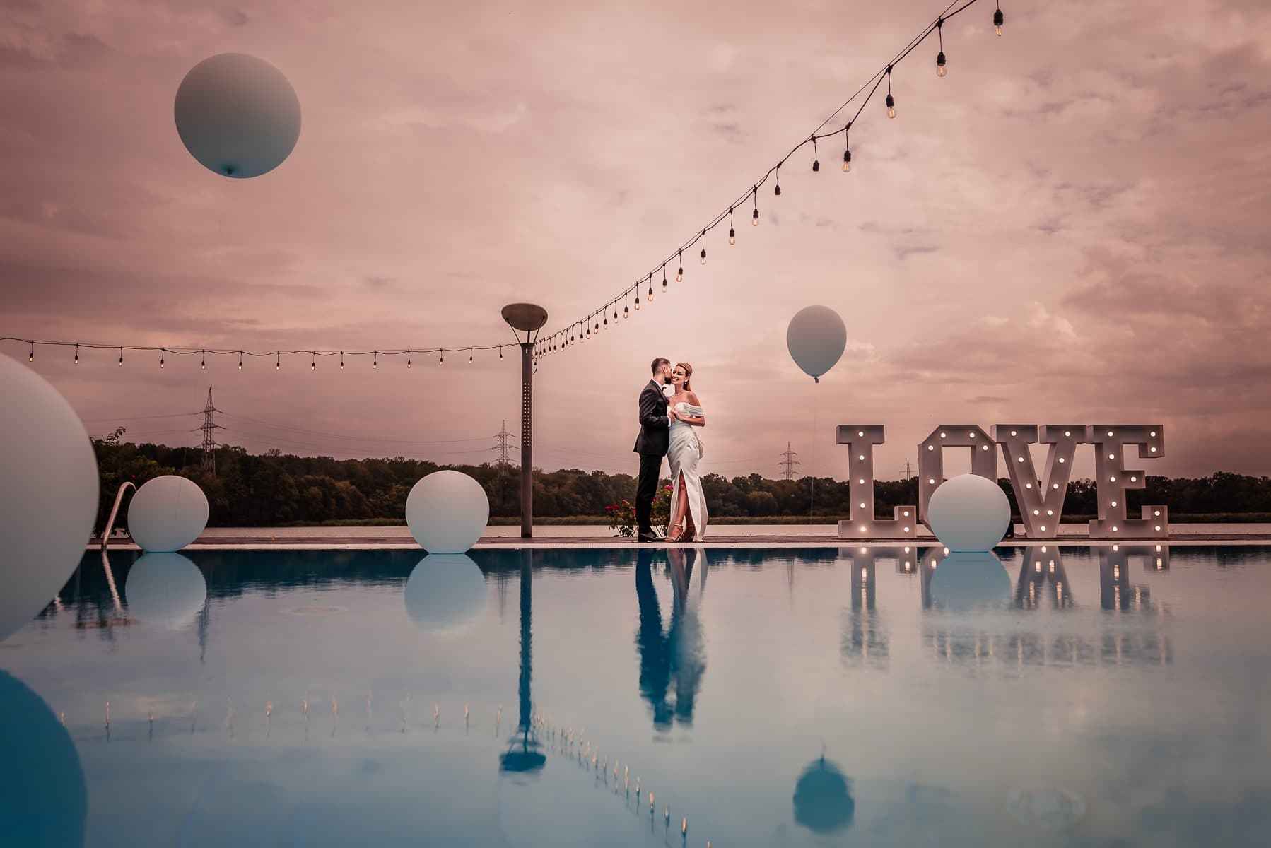 The couple reflected in a pool at dusk, balloons and LOVE letters on the deck