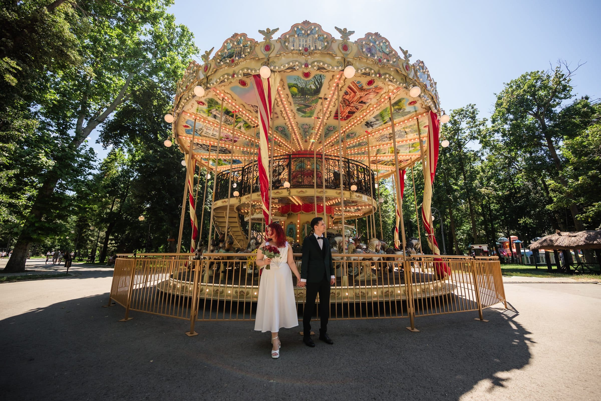Bride and groom standing small in front of a large vintage carousel in a sunny park