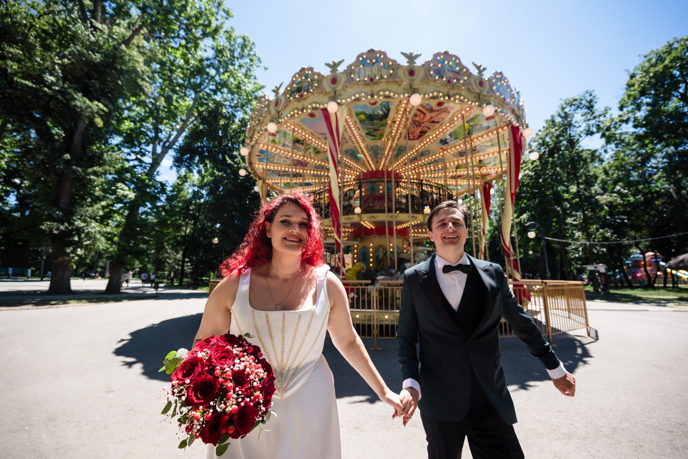 The bride with red hair and red roses walks hand in hand with the groom in front of the carousel