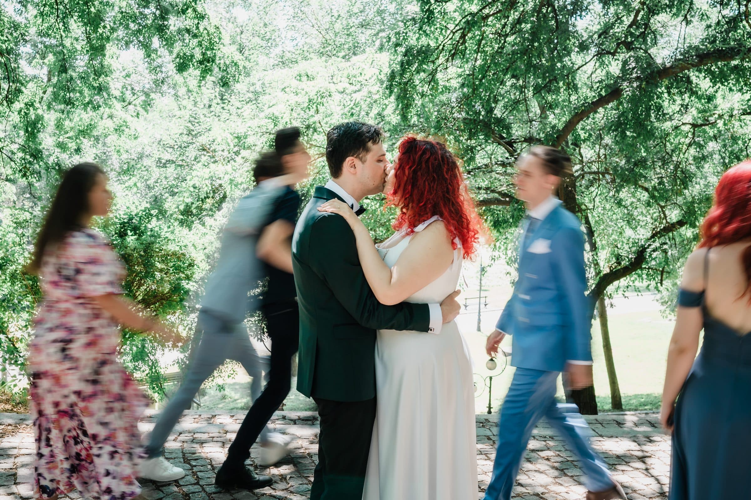 The couple kisses under the trees while guests walk past in a blur