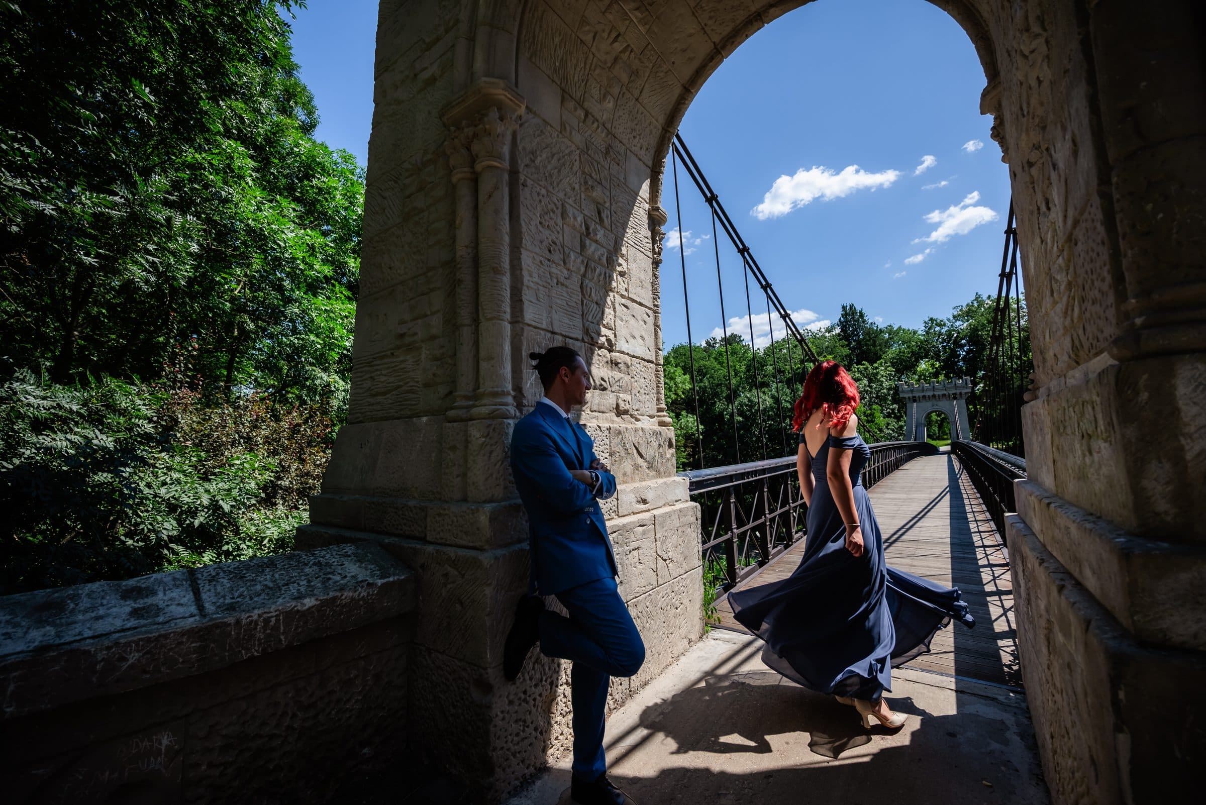 On a suspension bridge, the groom leans against a stone arch while the bride twirls in a blue dress