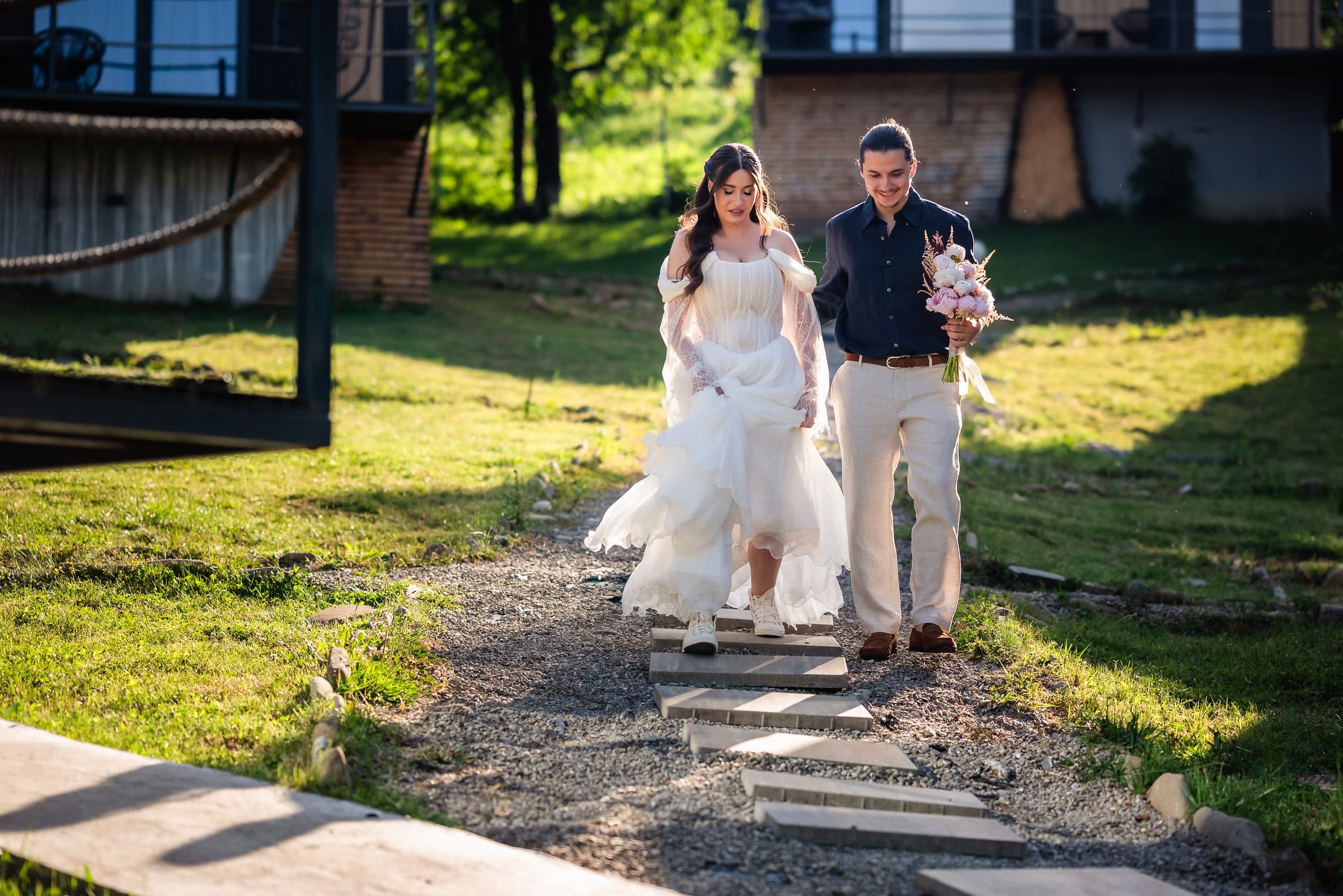 The bride walks down a stone path toward the ceremony, beside a man in a striped shirt
