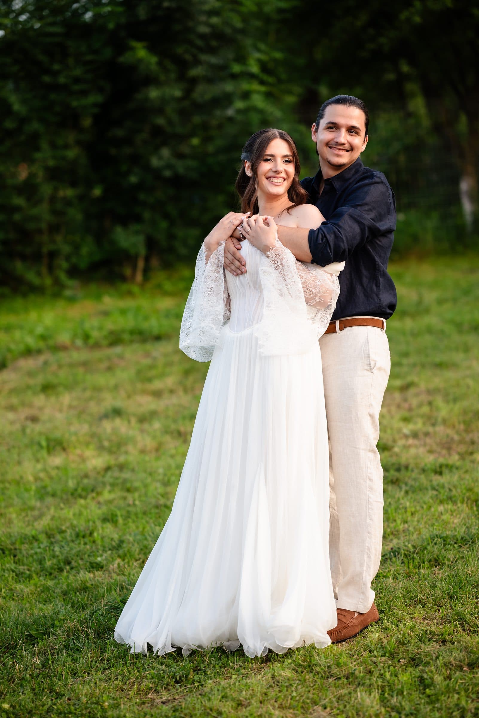 The groom wraps his arms around the bride from behind in evening light