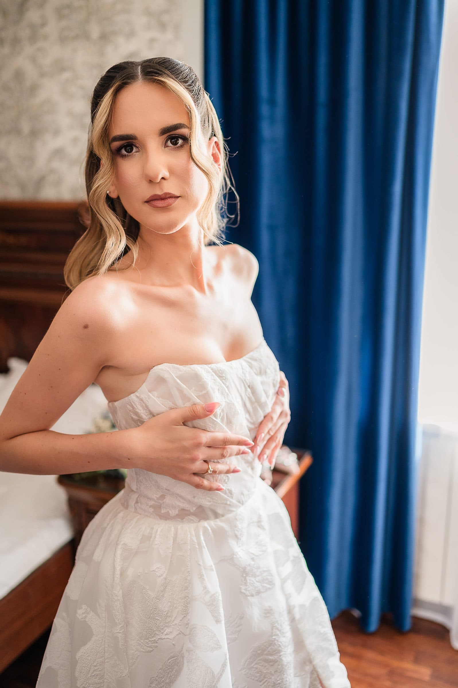 The bride stands by a blue curtain, hand on her dress