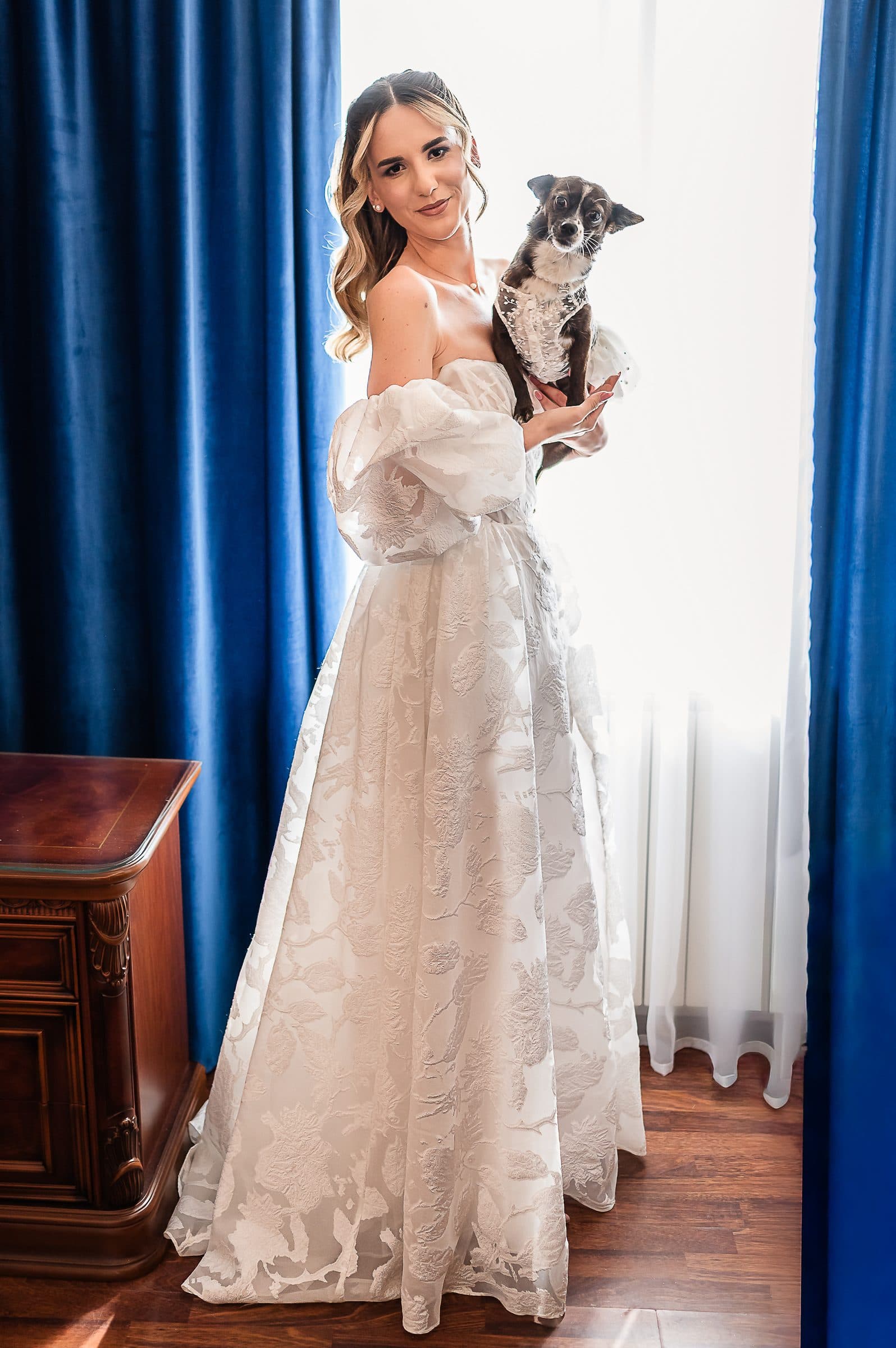 The bride holds the dog by the window, laughing