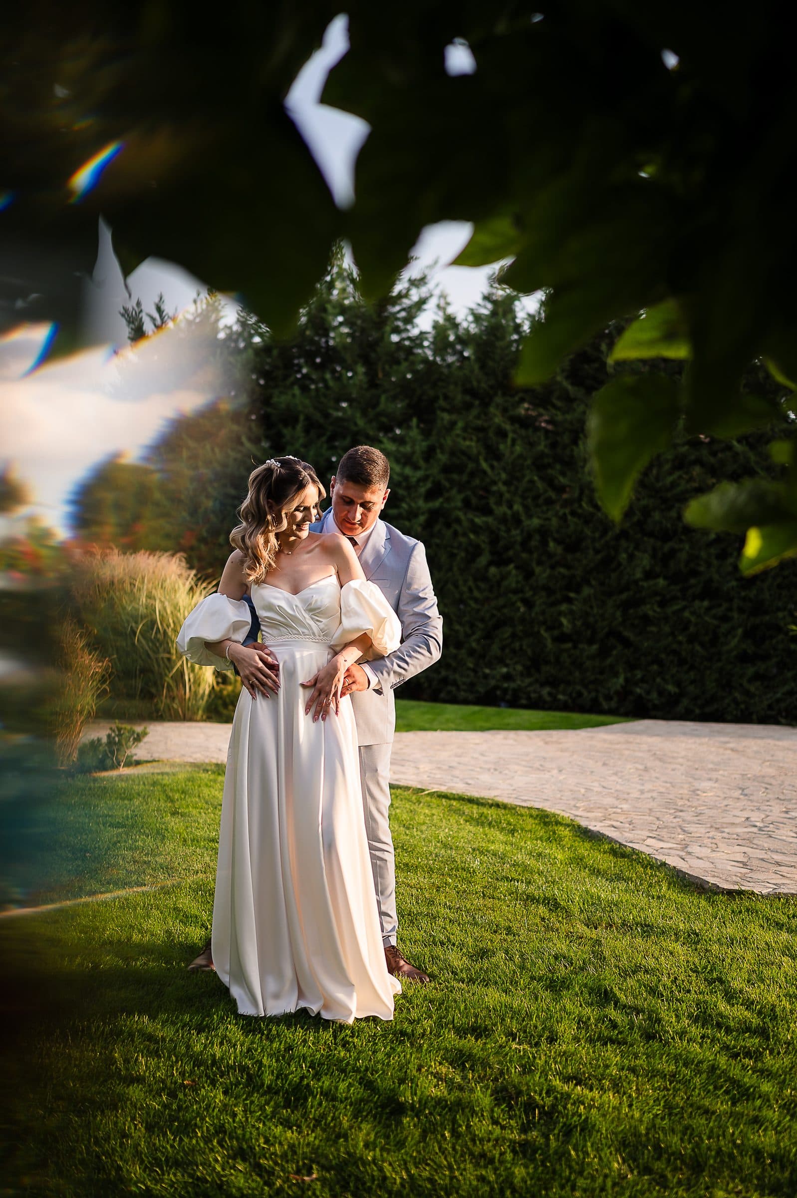 The couple stands close on the lawn, framed by leaves in the foreground