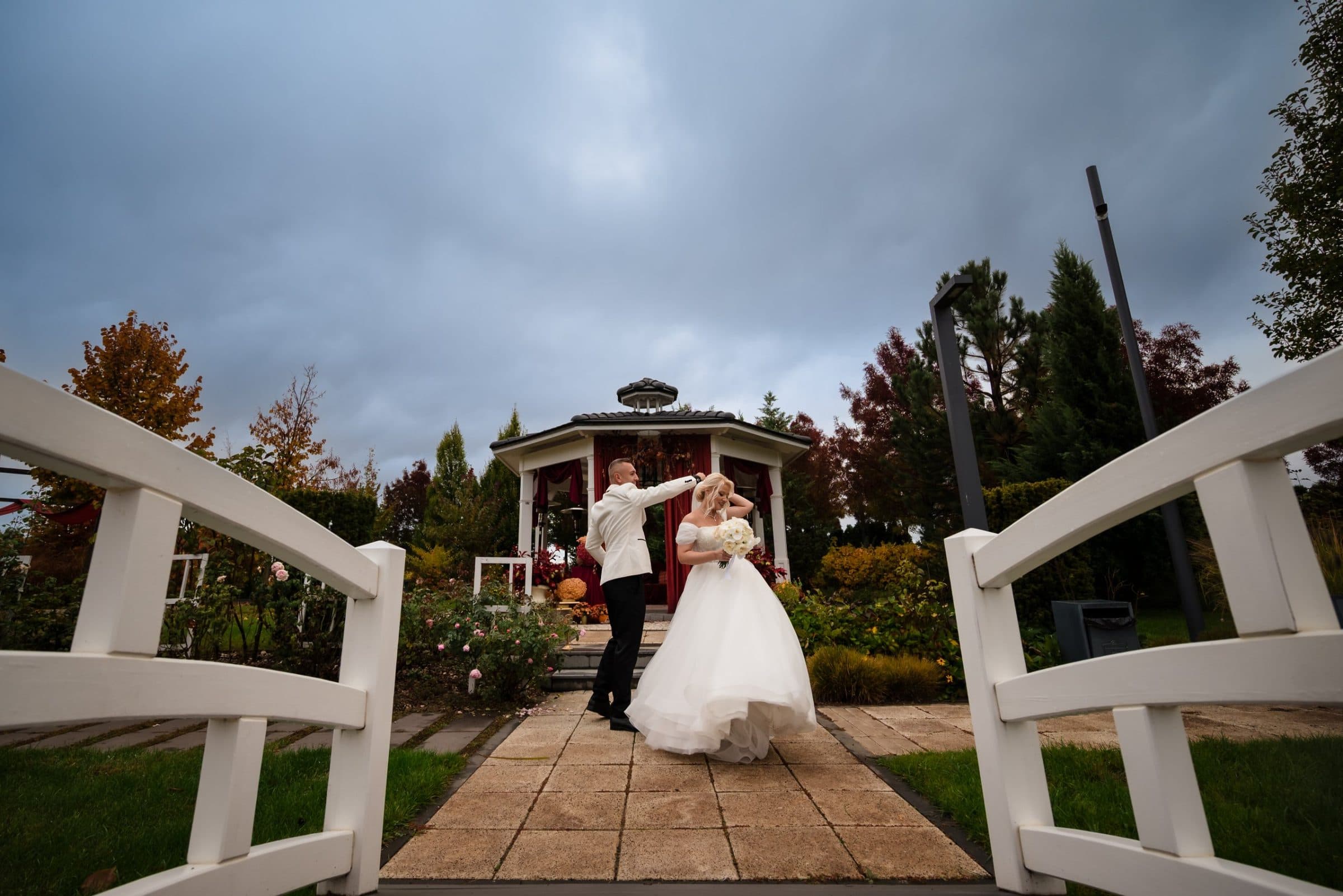 The groom twirls the bride in front of a garden gazebo, seen between white bridge railings