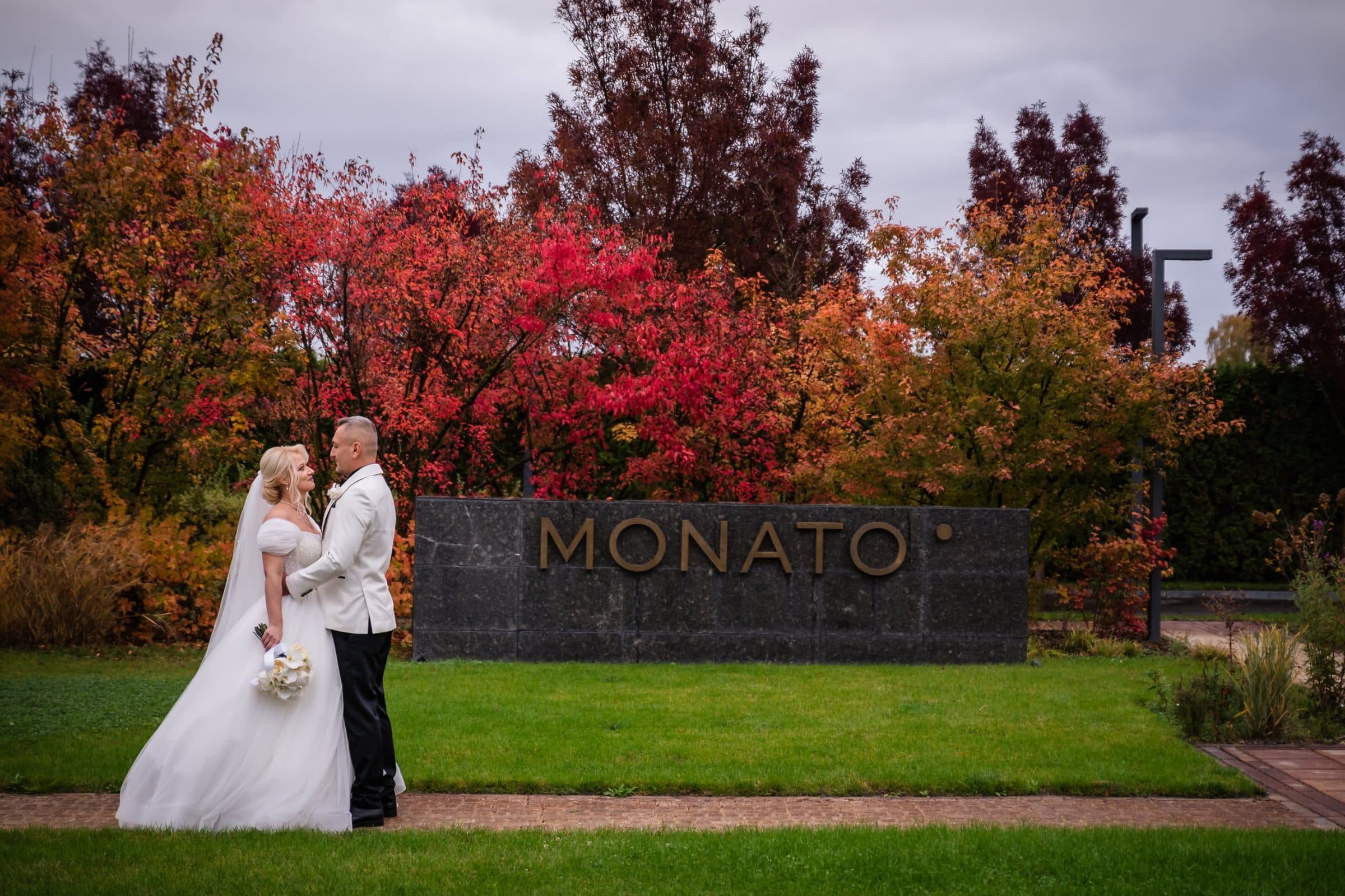 Bride and groom facing each other beside the Monato sign, under red and orange autumn trees