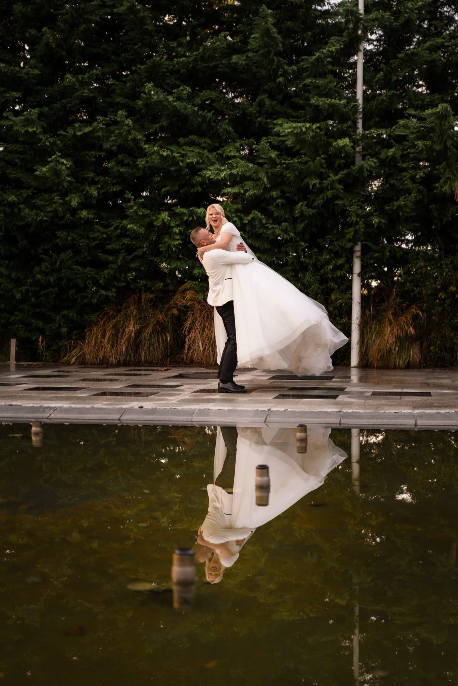 The groom lifts the laughing bride, reflected in a still pool