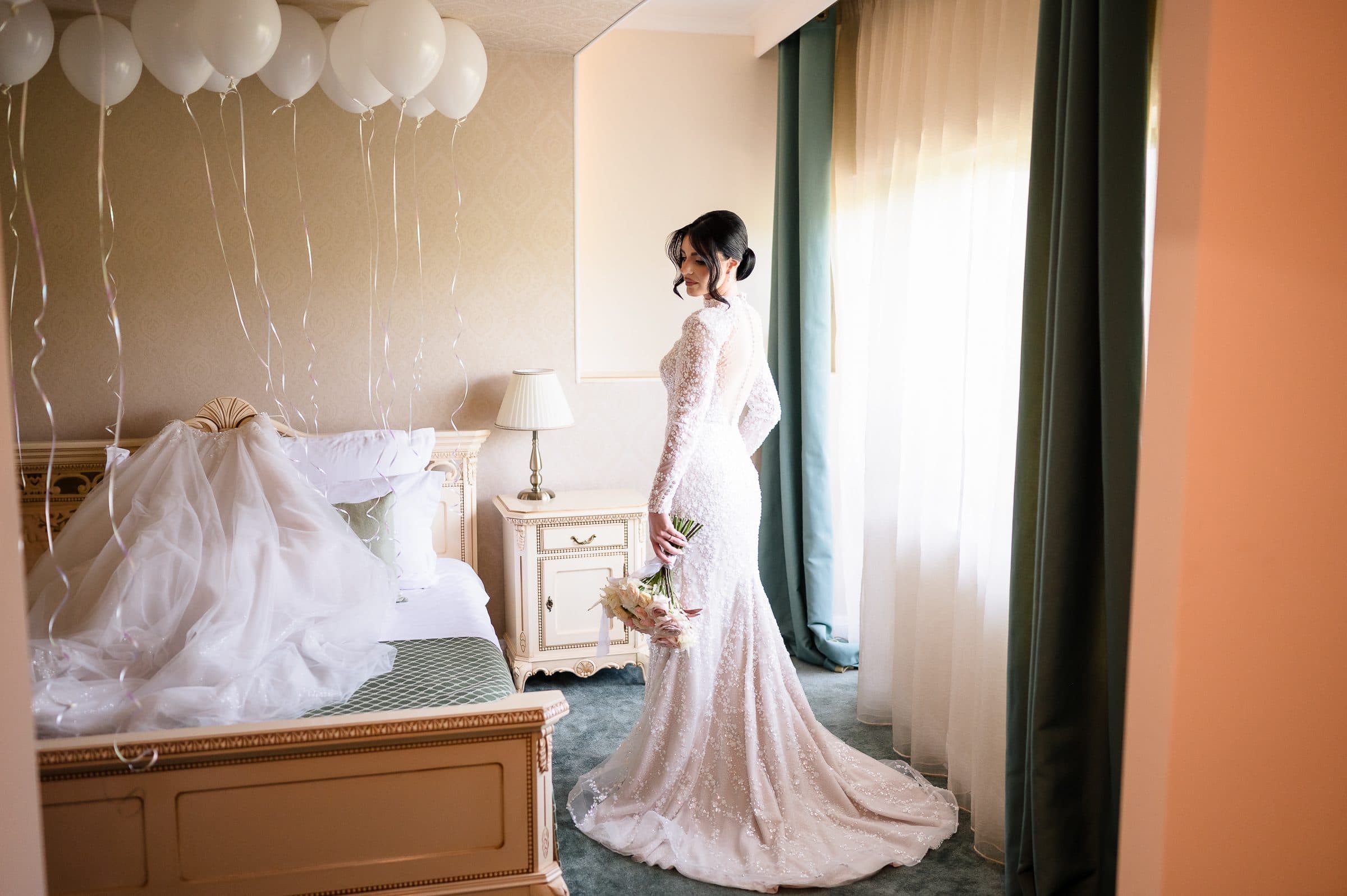 The bride stands by the window of her room, her veil spread across the bed beneath white balloons