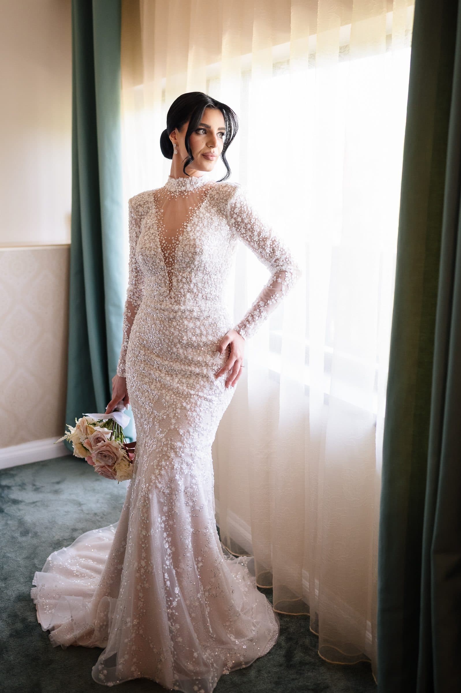 The bride in a beaded long-sleeved gown, standing in window light between green curtains