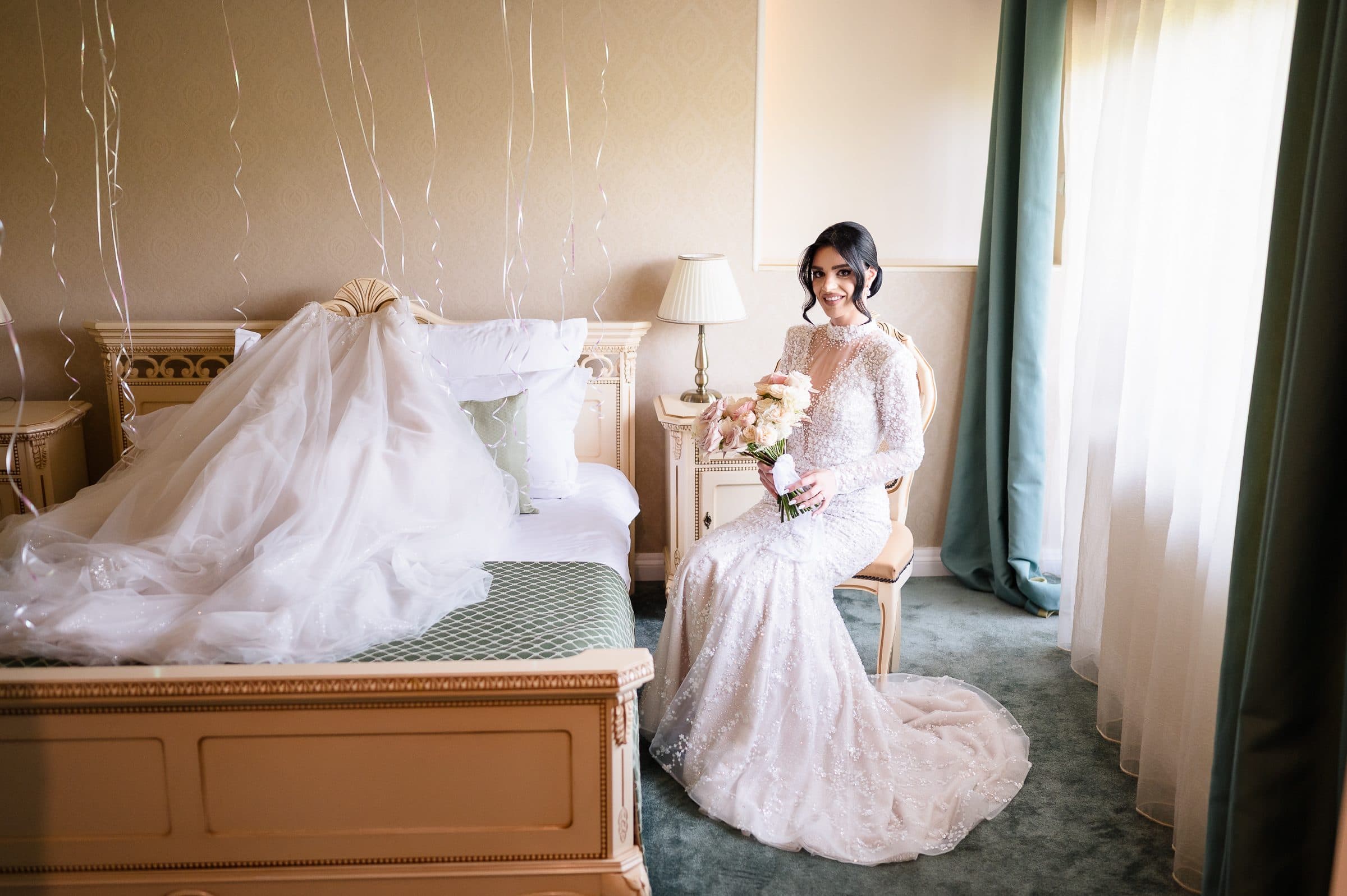 The bride smiles from a chair beside the bed where her veil is laid out