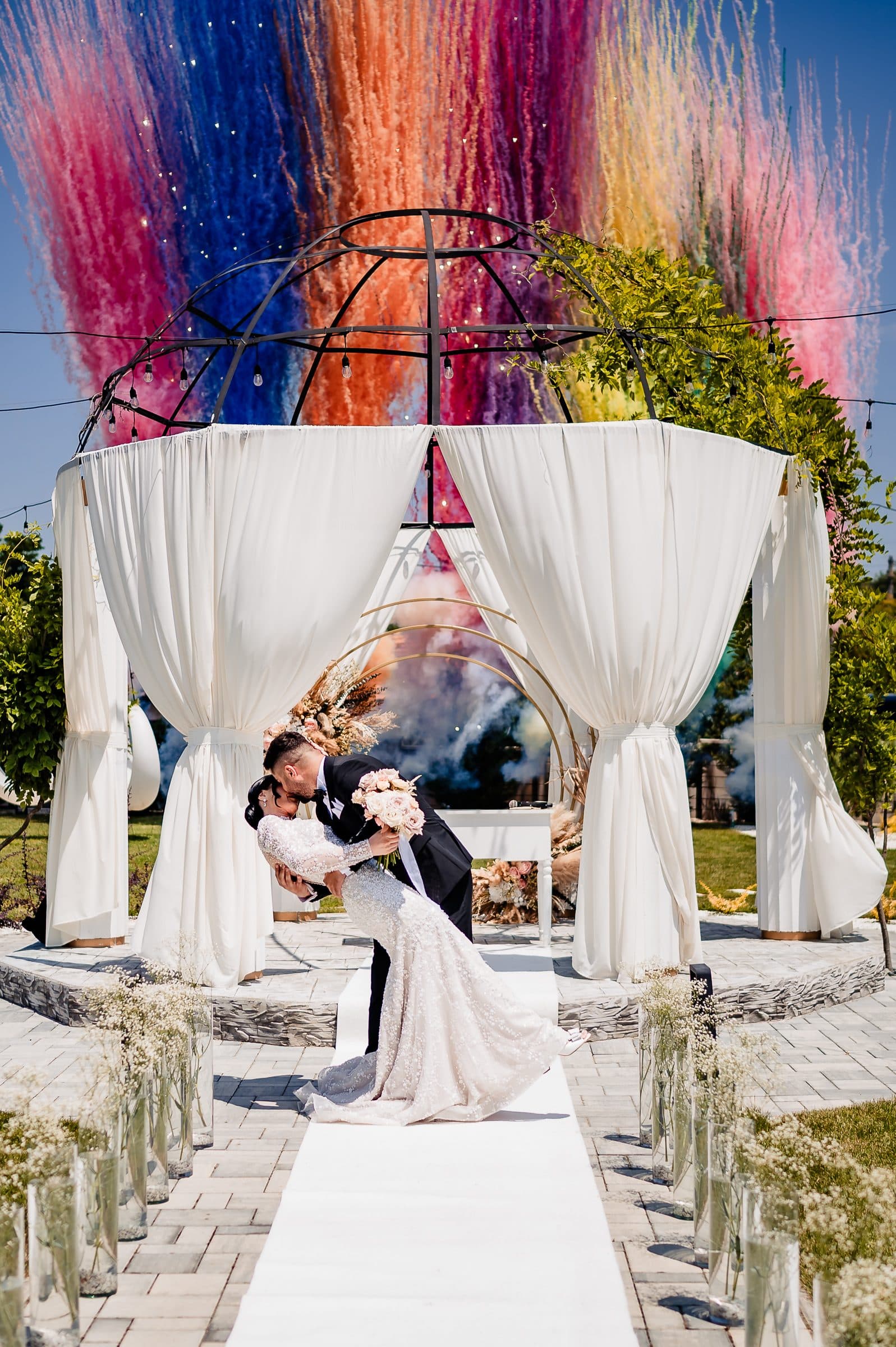 The groom dips the bride for a kiss in front of the ceremony pavilion as coloured smoke fills the sky