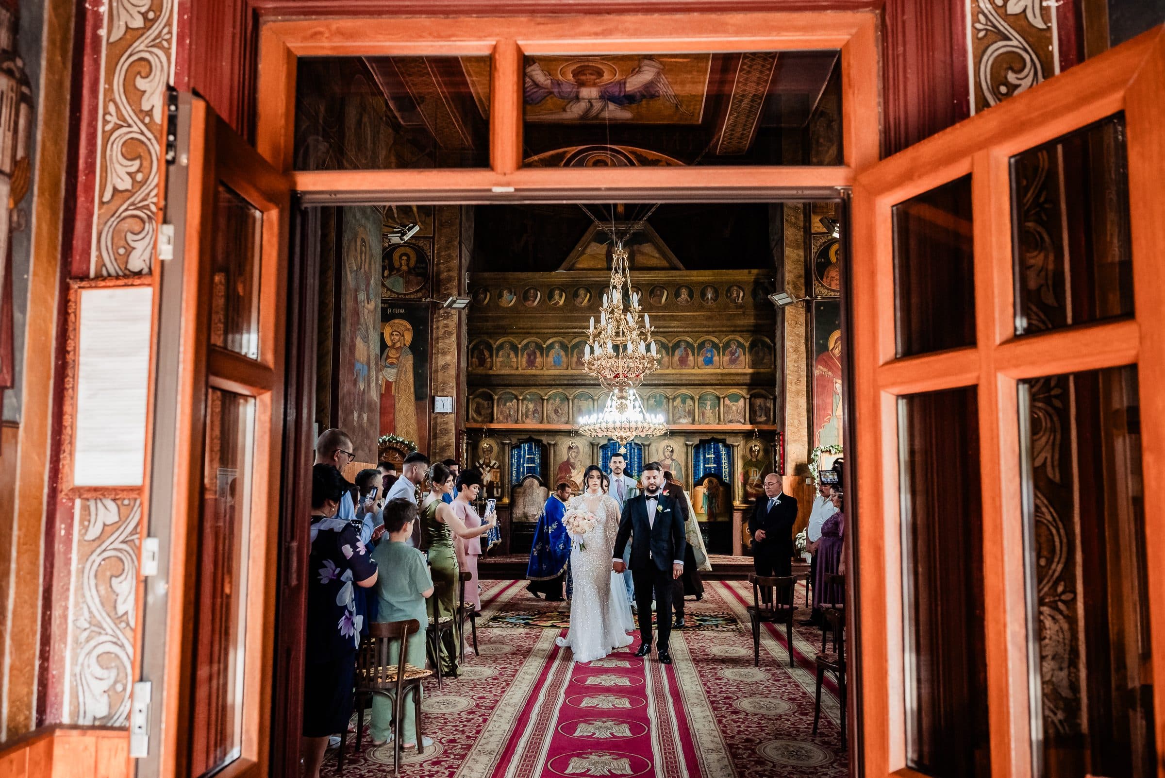 Seen through the open church doors: the couple walks the aisle under a chandelier while guests watch