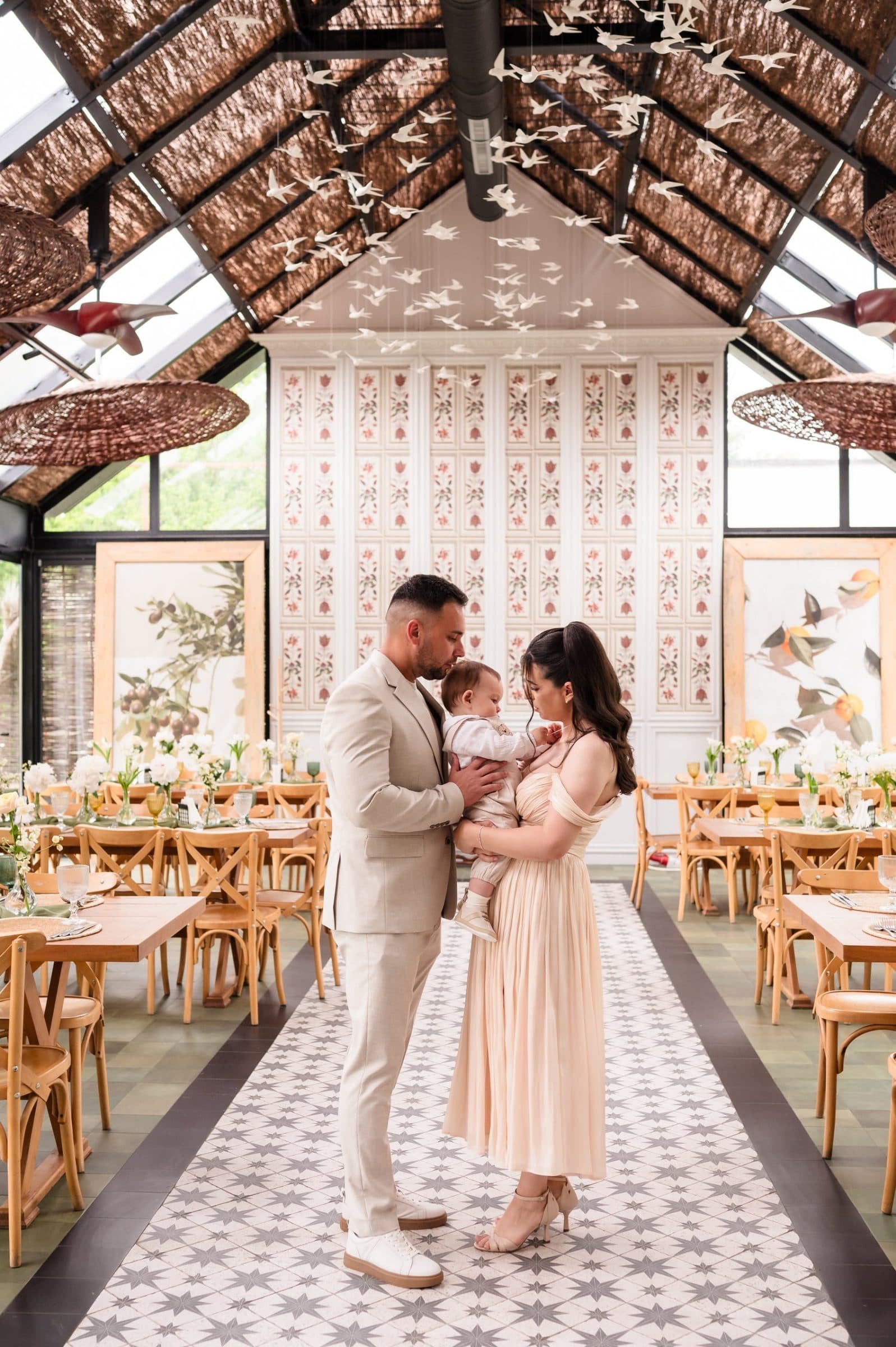 Parents holding their son in a glass-roofed hall, paper birds hanging above the set tables