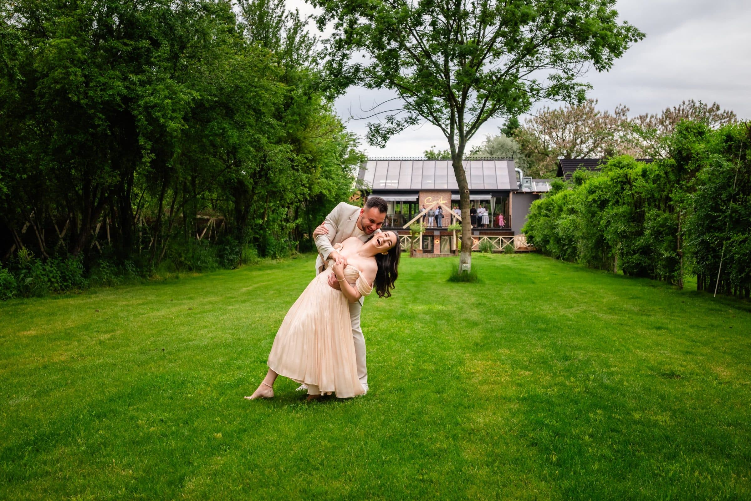 The father dips the laughing mother on the lawn while guests watch from the terrace